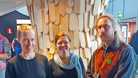 Kolme ihmistä hymyilee ryhmäkuvassa. Taustalla on puusta tehty taideteos.