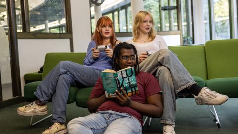 Nuoria kirjastossa. Etualalla poika lukemassa kirjaa. Takana olevalla sohvalla kaksi tyttöä pelaamassa pelikonsolilla.