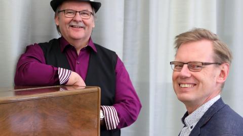 Man in a blue jacket is sitting by the piano, another man in a purple shirt is leaning on the piano further away. Both are smiling.