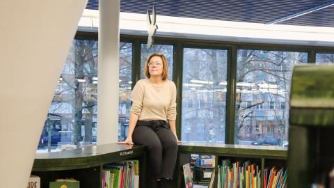 Nainen istuu ikkunan edessä kirjaston matalien kirjahyllyjen päällä ja hymyilee. 