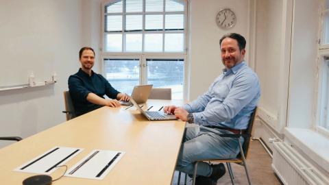 Kaksi miestä istuvat hymyillen kokouspöydän edessä läppärit kädessä. 