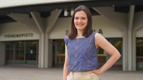 Henkilö seisoo rakennuksen edessä. Hän pitää kättään vyötäröllään.