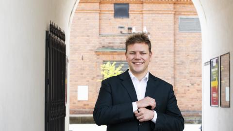 A person stands with their hands in front of them outside between white walls with a brick wall behind them.
