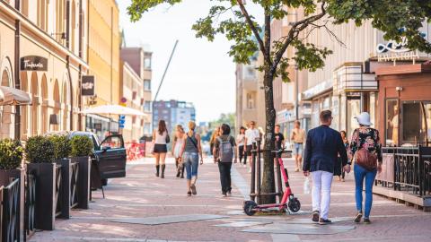 Ihmisiä kävelee kesäaikaan kadulla. Etualalla puu, johon nojaa sähköpotkulauta.