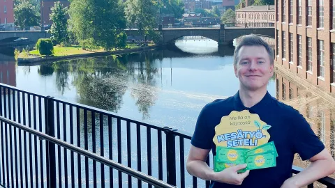 Petri Räsänen, the Summer Job Voucher Coordinator for the City of Tampere, is standing on Vapriikinraitti. He is holding the Summer Job Voucher logo and vouchers in his hand and smiling. In the background, you can see the city center of Tampere.