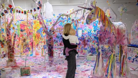 A woman stands with a child in her arms in a colorful room, where different colors have been painted on a white background. The woman and child are looking at a large unicorn made of paper.