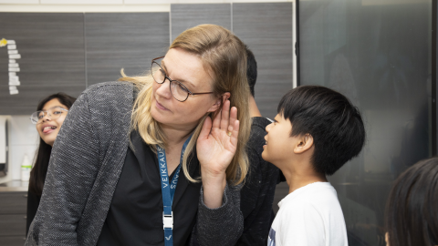 A child whispers in the teacher&#039;s ear. Another child in the background.