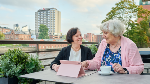 Kaksi ikääntynyttä henkilöä istuu pöydän ääressä edessään tablet-laite ja kahvikuppi.