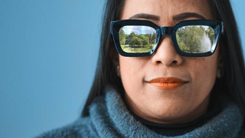 Tummahiuksinen henkilö katsoo suoraan kohti katsojaa. Hänellä on paksusankaiset aurinkolasit, joista heijastuu puiston kuva.