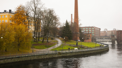 A landscape view featuring Tammerkoski, a red brick chimney, and the yellow Old Library building peeking out from behind the trees.
