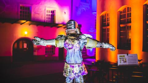 A metal statue of a human with their arms outstretched. The statue is wearing a helmet. In the background of the work you can see walls illuminated by colourful lights.