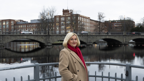 A person is standing in front of a metal railing. Behind them, you can see the rapids, the Satakunnansilta bridge with cars, and the Tampella buildings.