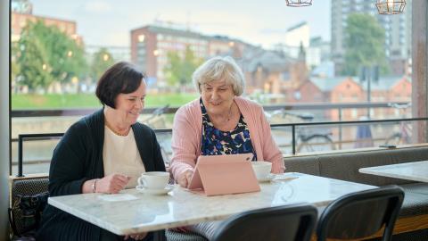 Two people are sitting in a café, smiling as they look at a tablet device.