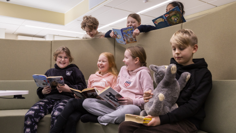 Koulun oppilaita istuu kirjojen ja pehmolelun kanssa ja nauraa. Osa oppilaista kurkkii ylhäältä sermien takaa.