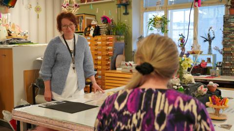 Henkilö seisoo pöydän ääressä. Etualalla toinen henkilö, joka tekee käsitöitä.