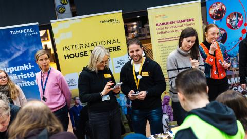 Several smiling city employees at information booths in Tampere Residents' Evening. Behind them are colorful roll-ups.