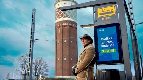 Cowboy-hattuinen mies nojaa pysäkkikatokseen, jossa näkyy mainos: Ratikkalinjasto laajenee 7.1.2025.