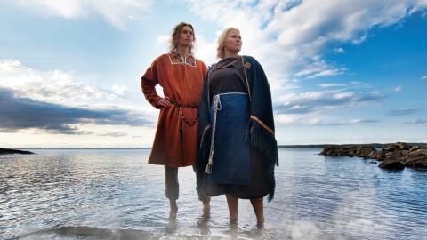 Mies ja nainen seisovat avojaloin matalassa vedessä, josta heijastuu sininen taivas. Pari on pukeutunut muinasipukuihin, naisella on sininen asu ja miehellä oranssi..