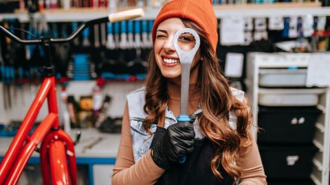 A long‑haired young person wearing a beanie peers through the hole of a large spanner. A bicycle repair workshop with tools is visible in the background.