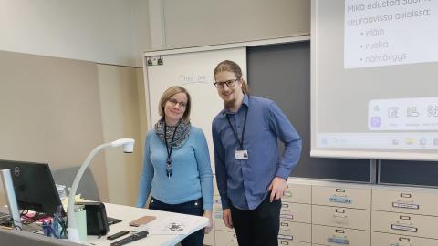 Two people are standing in a classroom by the teacher’s desk.