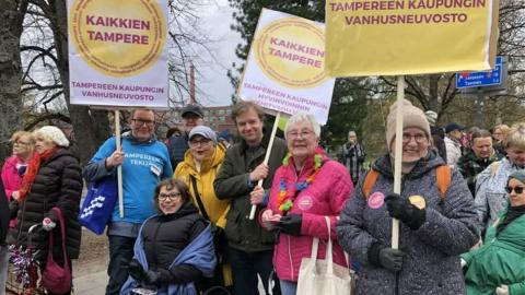 Hymyileviä ihmisiä mielenosoituskylttejä käsissään. Niissä lukee Kaikkien Tampere ja Tampereen kaupungin vanhusneuvosto.