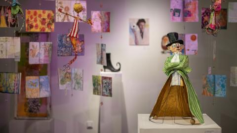 Kuva galleriasta, jossa etualalla lierihattupäinen veistos ja taka-alalla on katosta roikkuvia värikkäitä teoksia. 