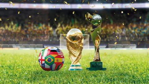 Two trophies and a soccer ball on a soccer field. People in the stands in the background.