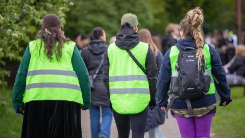 Kolme henkilöä kävelee rinnakkain vihreät huomioliivit yllään ihmisten keskellä.