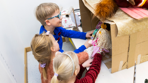Three children play puppet theatre holding two puppets inside a cardboard box.