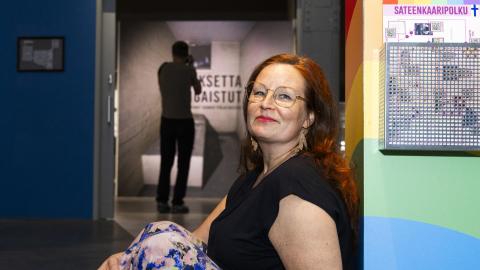 A red-haired woman in a black short-sleeved blouse is leaning against the wall. In the background, a museum display case stands out.