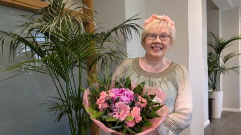 Aija Karttunen, with her light hair and a smile on her face, is standing next to a potted plant. She is holding a large bouquet of light pink flowers.