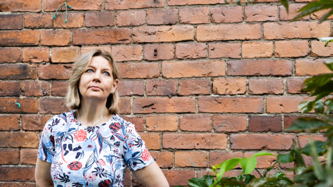 A person wearing a floral shirt is standing in front of a brick wall. Next to them are leaves from a bush.