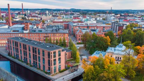 Ilmakuva Tampereen keskustasta, jossa näkyvät Tammerkosken punatiiliset tehdasrakennukset, syksyinen puistoalue ja kaupungin siluetti.