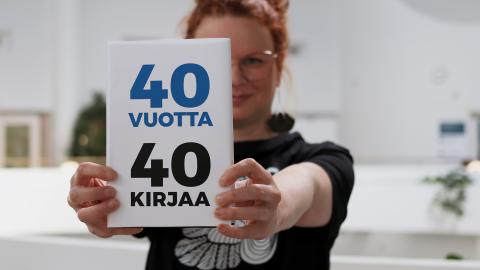 Tuija seisoo Metson parvikerroksessa ja pitää osittain kasvojensa edessä kirjaa, jonka kannessa on teksti 40 vuotta, 40 kirjaa. 