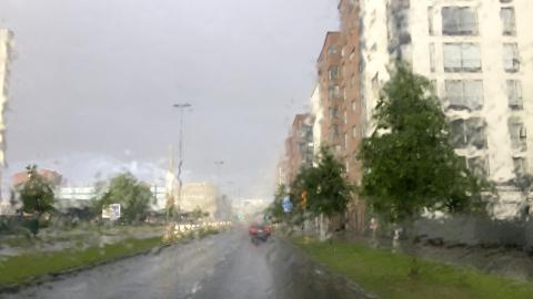 Rankkasateen kastelema kaupunkikatu auton tuulilasin läpi nähtynä; märkä ajorata, liikennettä ja kerrostaloja tumman sadepilven alla.