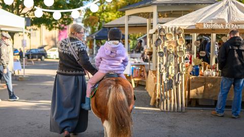 A woman is leading a pony, with a child sitting on its back, wearing a riding helmet.