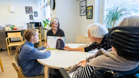 Lapsi näyttää vanhukselle kirjasta lukemaansa sivua. Opettaja ja toinen vanhus seuraavat.