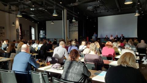 Sali on täynnä osallistujia istumassa pöytien ääressä ja seuraamassa esiintymislavalla käytävää paneelikeskustelua.