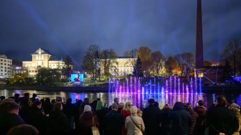 Yleisö seuraa iltahämärässä Tammerkosken rannalla Tanssivia vesiä. Veden pinnasta nousevat sinisin ja violetin sävyin valaistut suihkulähteet, taustalla näkyy valaistuja rakennuksia, tehdaspiippu sekä taivaalle suuntautuvia valokeiloja.