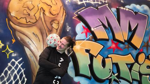 Mies temppuilee jalkapallon kanssa. Taustalla värikäs graffiti maalaus, jossa teksti MM Futis.