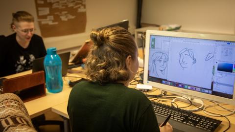 Kaksi henkilöä istuu työpöytien äärellä. Toisen tietokoneen ruutu näkyy kameralle, ja ruudulla on piirroksia.