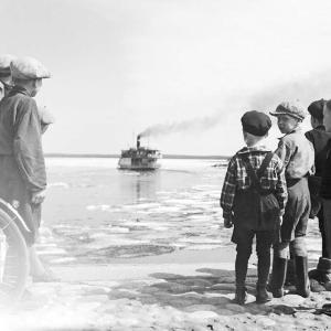 Järvellä höyrylaiva, lapsia seisoo rannalla katsomassa laivaa. Mustavalkoinen kuva.