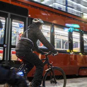 Aikuinen ja lapsi pyöräilevät talvella kadulla. Heillä on heijastimet, valot ja kypärät. Raitiovaunu ohittaa heidät. 