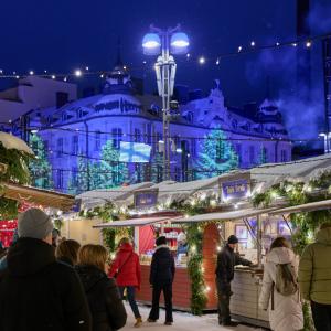 Ihmisiä hämärän aikaan Joulutorilla joulukoristelujen ja valaistujen puotimökkien äärellä, taustalla valotaiteella valaistu rakennus.