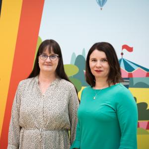Tampereen kaupungin lapsiasiamiehet Tiia Heinäsuo ja Taru Kuosmanen seisovat rinnakkain. Heidän takanaan on värikäs, voimakkaasti kuvioitu seinä, jossa erottuu karkkeja ja sirkusteltta.