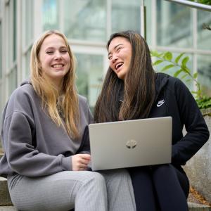 Kaksi nuorta naista istuvat yliopistokampuksen ulkorappusilla tietokoneen kanssa ja nauravat.