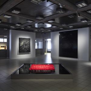 View from the ground floor of the museum, dimly lit. Huge red and white glass beads hang from the ceiling, behind which is a landscape window looking out onto a snow-covered sculpture park. In the centre of the picture is a large pool of ink with red roses in the middle. Behind it are two white partitions, one with a black non-representational relief hanging on it, the other with a black painting almost as high as the partition with representational elements. Both the relief and the painting are reflected i