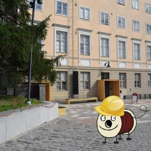 Taustalla vaalea vanha kivirakennus, jonka edessä mukulakivipiha. Edustalla piirretty Rulla-hahmo seisoo päässään keltainen rakennuskypärä.