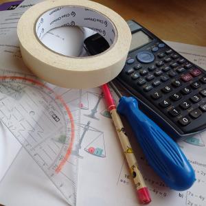 Matematiikan kirjan päällä laskin, maalarinteippiä, ruuvimeisseli, kynä ja piirtokolmio.
