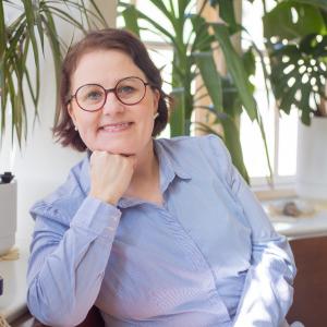 Uusi nuorisopalvelujohtaja Tiina-Liisa Vehkalahti katsoo kameraan hymyillen. Ympärillä huonekasveja ja vanhanaikainen kirjoituskone.